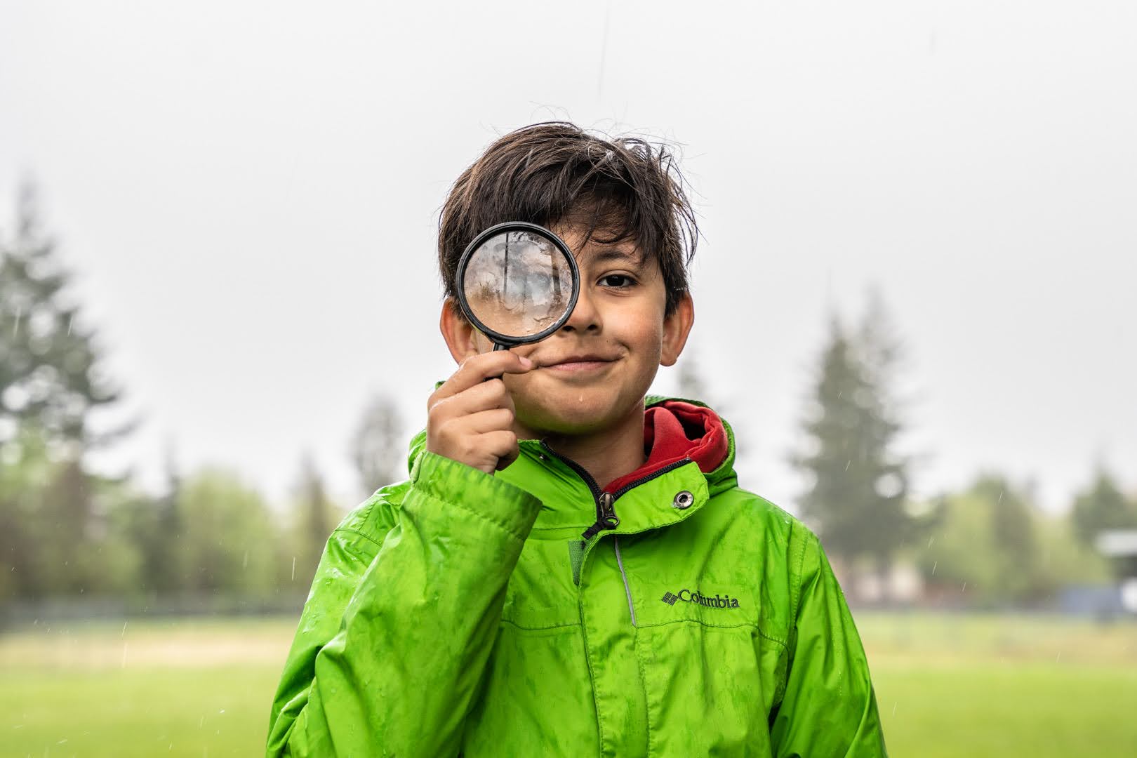 A boy holds a magnifying glass up with a forest in the background.