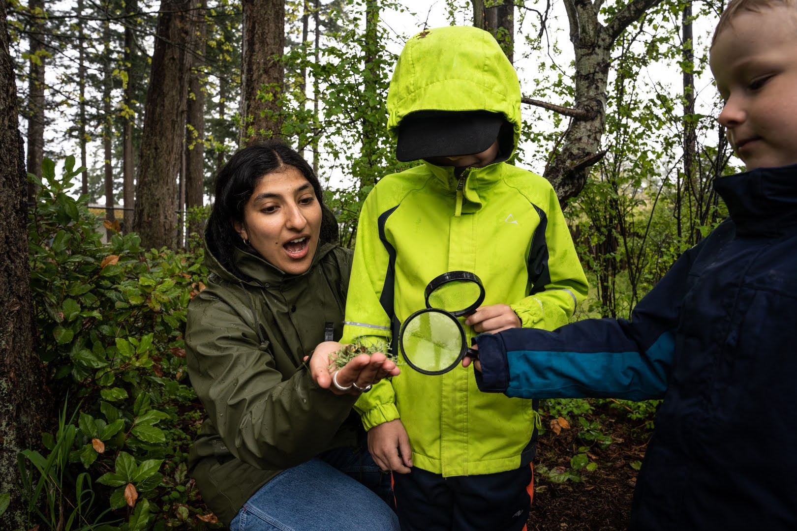 An environmental educator leads a school workshop, showing two students with magnifying glasses some lichen.